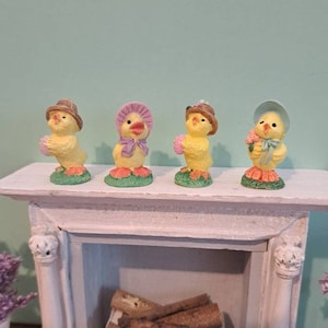 May include: Four yellow chicks wearing hats and holding flowers are arranged on a white mantelpiece above a fireplace. The fireplace has a white mantel and a white hearth with a white lace doily and a pile of logs.