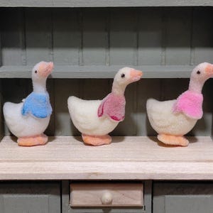 May include: Three fluffy white goose figurines with pink, blue, and pink scarves. The geese have orange feet and beaks. They are displayed on a wooden shelf inside a miniature cabinet.
