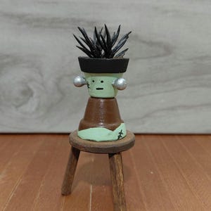 May include: A handcrafted, painted miniature Frankenstein-themed flower pot with a small succulent plant. The pot is brown and green, with silver accents, and sits on a small wooden stool. The plant has dark green spiky leaves.
