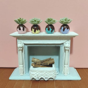 May include: Miniature fireplace with four succulent plants in colorful pots. The fireplace is painted white and light blue, with a decorative lace basket holding wooden logs. The succulent pots are pink, yellow, teal, and purple.