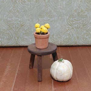 May include: A miniature terracotta pot with yellow flowers sits on a small wooden stool. A small white pumpkin with a green stem is next to the stool. The scene is set on a wooden surface.
