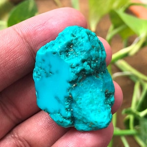 May include: A raw, unpolished turquoise gemstone with a vibrant, saturated cyan-blue colour. The stone features an irregular, textured surface with natural crevices and small dark inclusions, showcasing a mix of smooth and rough areas.