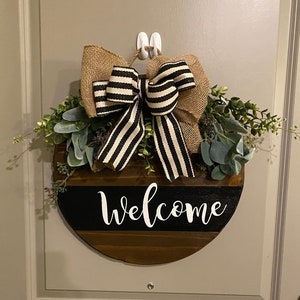 Puede incluir: Un letrero de bienvenida redondo de madera con un lazo a rayas blanco y negro y vegetación. El letrero dice "bienvenido" en letras blancas.