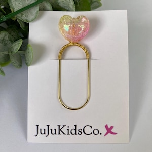 May include: A heart-shaped bookmark with a gold-coloured wire design. The heart is filled with iridescent glitter in shades of pink, yellow, and orange. The bookmark is attached to a white card with the text "JujuKidsCo." and a pink "X" logo.