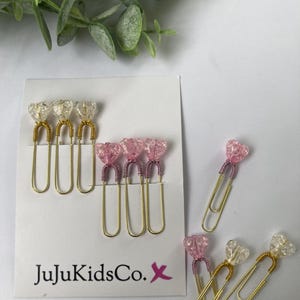 May include: A collection of decorative paper clips featuring heart-shaped beads in clear and pink hues. The beads are attached to gold-tone wire clips with coiled wire accents. A white display card with the text JuJuKidsCo. X is included.