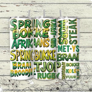 May include: Three white stainless steel tumblers with green and yellow text. The text includes the words "SPRINGBOKKE", "AFRIKAANS", "BRAAI", and "STEAK". The tumblers have black straws and are on a white wooden background.