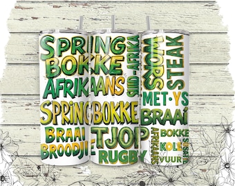 May include: Three white stainless steel tumblers with green and yellow text. The text includes the words "SPRINGBOKKE", "AFRIKAANS", "BRAAI", and "STEAK". The tumblers have black straws and are on a white wooden background.
