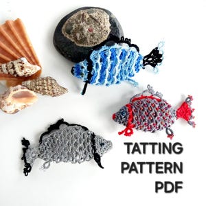 以下が含まれることがあります： 青、グレー、赤の3匹の手作りのタティングレースの魚、黒のアクセント付き。 魚は貝殻と石と一緒に白い背景に配置されています。 テキスト「TATTTING PATTERN PDF」も表示されています。
