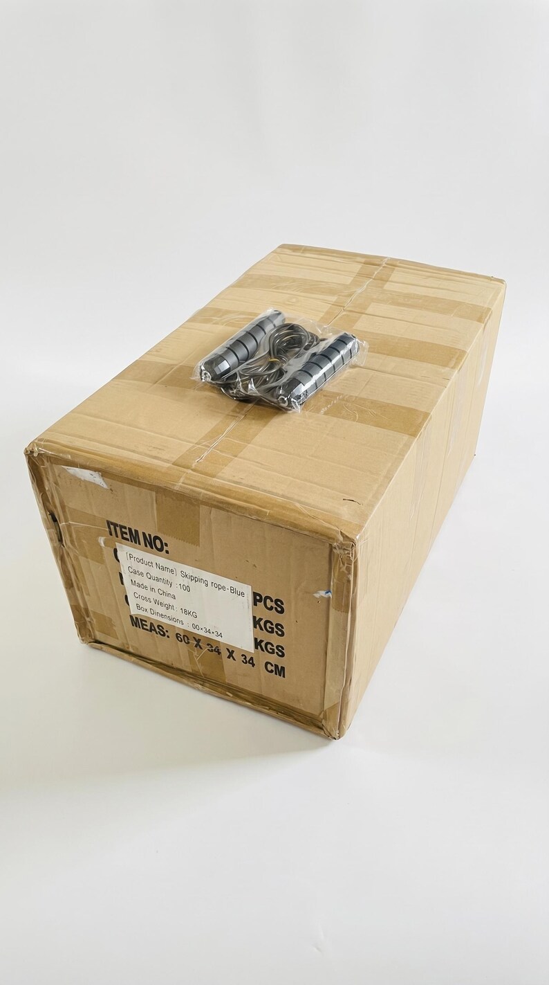 May include: A brown cardboard shipping box with a skipping rope in clear plastic packaging on top. The box features a printed label with the text: ITEM NO: Product Name: Skipping rope-Blue, Case Quantity: 100 PCS, Made in China, Cross Weight: 18KG, Box Dimensions: 60x34x34, MEAS: 60 X 34 X 34 CM.