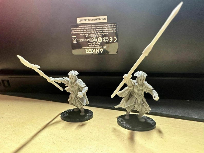 K&ouml;nnte beinhalten: Zwei wei&szlig;e Plastikfiguren von Elfen, die Speere halten. Die Elfen tragen R&uuml;stungen und Umh&auml;nge. Die Figuren stehen auf schwarzen runden Sockeln.