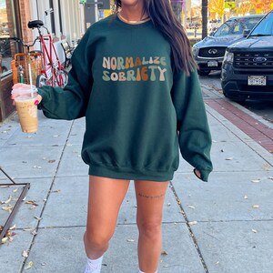May include: A dark green sweatshirt with the text "NORMALIZE SOBRIETY" in rainbow colours. The sweatshirt is being worn by a person with white trainers and white socks.