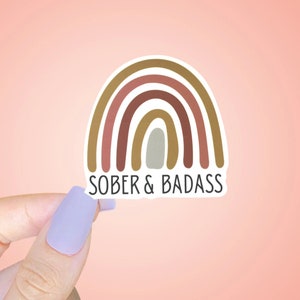 May include: A sticker with a rainbow design in shades of brown, pink, and gray. The text "SOBER & BADASS" is printed below the rainbow.