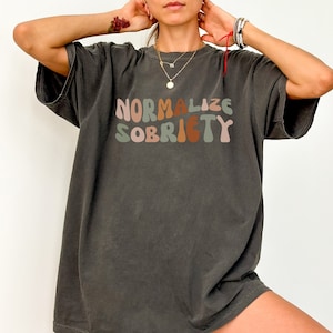 Puede incluir: Una camiseta gris oscuro de gran tamaño con el texto "Normalizar la sobriedad" impreso en letras coloridas y onduladas.