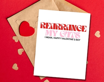 May include: A white card with the text "Rearrange My Guts I mean, Happy Valentine's Day" in red and pink letters. The card is sitting on a brown envelope with red hearts scattered around it.