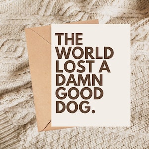 May include: A greeting card with the text "THE WORLD LOST A DAMN GOOD DOG" in dark brown letters on an off-white background. The card is paired with a brown envelope, set against a textured, cream-colored surface.