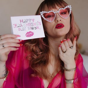 May include: A pink card with the text "Happy Galentines Day Bitch" in pink and white letters with a pink lip print below. A person is holding the card in front of their face, wearing pink heart-shaped sunglasses with pearl accents.