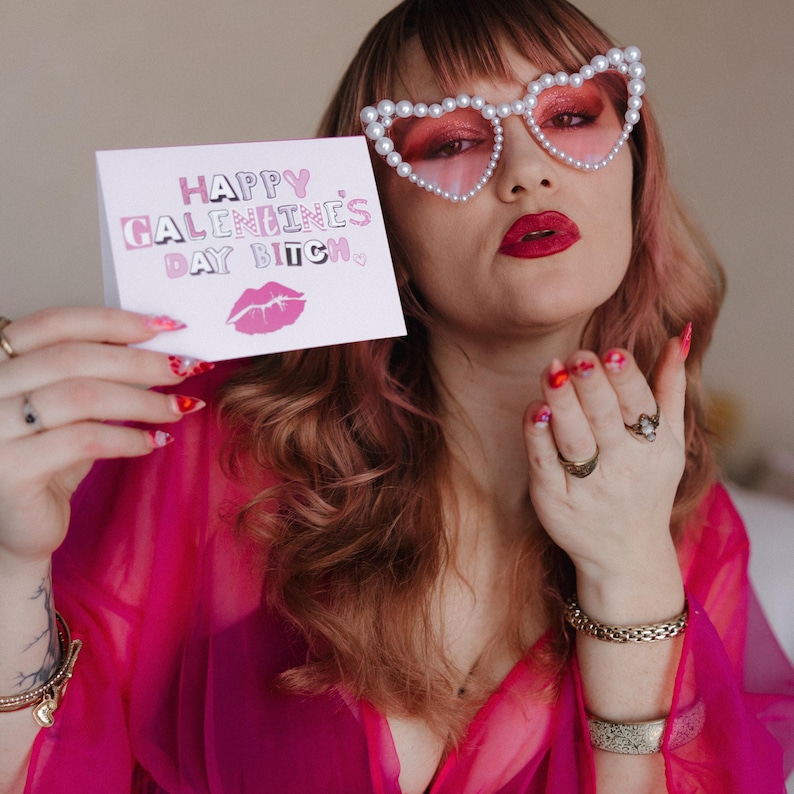 May include: A pink card with the text "Happy Galentines Day Bitch" in pink and white letters with a pink lip print below. A person is holding the card in front of their face, wearing pink heart-shaped sunglasses with pearl accents.