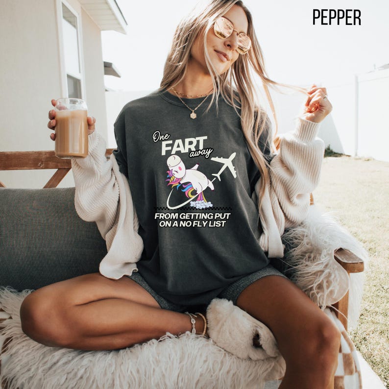 Op de afbeelding: Een antracietgrijs T-shirt met korte mouwen en een afbeelding van een eenhoorn met een regenboogkleurige manen en staart die naast een vliegtuig vliegt. De tekst luidt One FART away FROM GETTING PUT ON A NO FLY LIST. Het shirt heeft een ronde hals.
