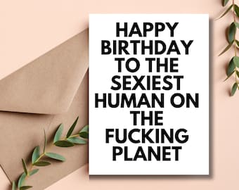 May include: A white greeting card with the text "HAPPY BIRTHDAY TO THE SEXIEST HUMAN ON THE FUCKING PLANET" in bold black letters. The card is next to a brown envelope on a light pink surface, with green leaves in the background.