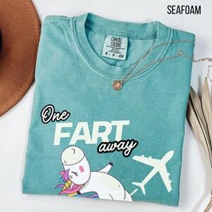Op de afbeelding: Een zeegroen T-shirt met ronde hals en een afbeelding van een witte eenhoorn met kleurrijke manen en een wit vliegtuig. De tekst luidt One FART away. Het shirt heeft een Comfort Colors merklabel in de nek en een gouden ketting.