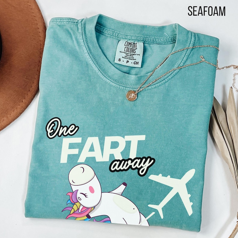 Op de afbeelding: Een zeegroen T-shirt met ronde hals en een afbeelding van een witte eenhoorn met kleurrijke manen en een wit vliegtuig. De tekst luidt One FART away. Het shirt heeft een Comfort Colors merklabel in de nek en een gouden ketting.