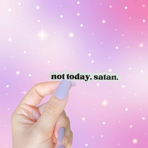 May include: A hand holding a white sticker with the text "not today, satan." The sticker is against a pink and purple background with white dots.