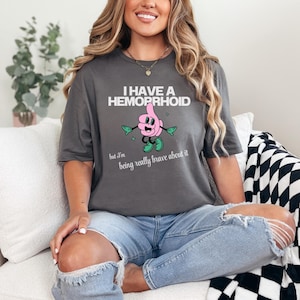 Puede incluir: Camiseta gris oscuro con el texto "I HAVE A HEMORRHOID" encima de un personaje de dibujos animados con el pulgar hacia arriba. El personaje es rosa con brazos y piernas verdes. Debajo, el texto dice "but I'm being really brave about it."