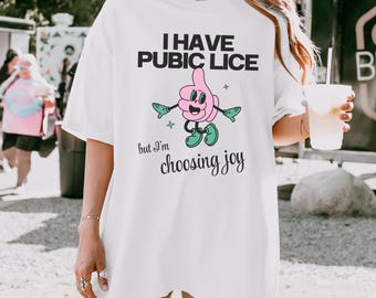 May include: A white t-shirt featuring a graphic of a pink cartoon character with green shoes and gloves, accompanied by the black text "I HAVE PUBIC LICE but I'm choosing joy" printed in a mix of bold block letters and cursive script.