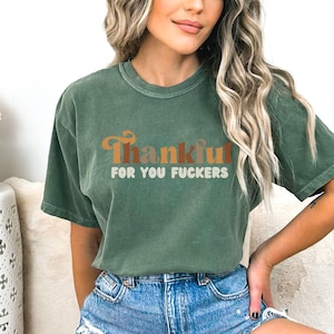 Op de afbeelding: Een bosgroen t-shirt met korte mouwen en de tekst Thankful in een groot, oranje, cursief retro-lettertype, gevolgd door de woorden FOR YOU FUCKERS in een kleiner, wit, afgerond schreefloos lettertype in het midden op de borst.