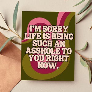 May include: A rectangular greeting card featuring the text I'M SORRY LIFE IS BEING SUCH AN ASSHOLE TO YOU RIGHT NOW. The design includes wavy, overlapping bands of pink, dark red, and olive green colors against a dark olive green background.