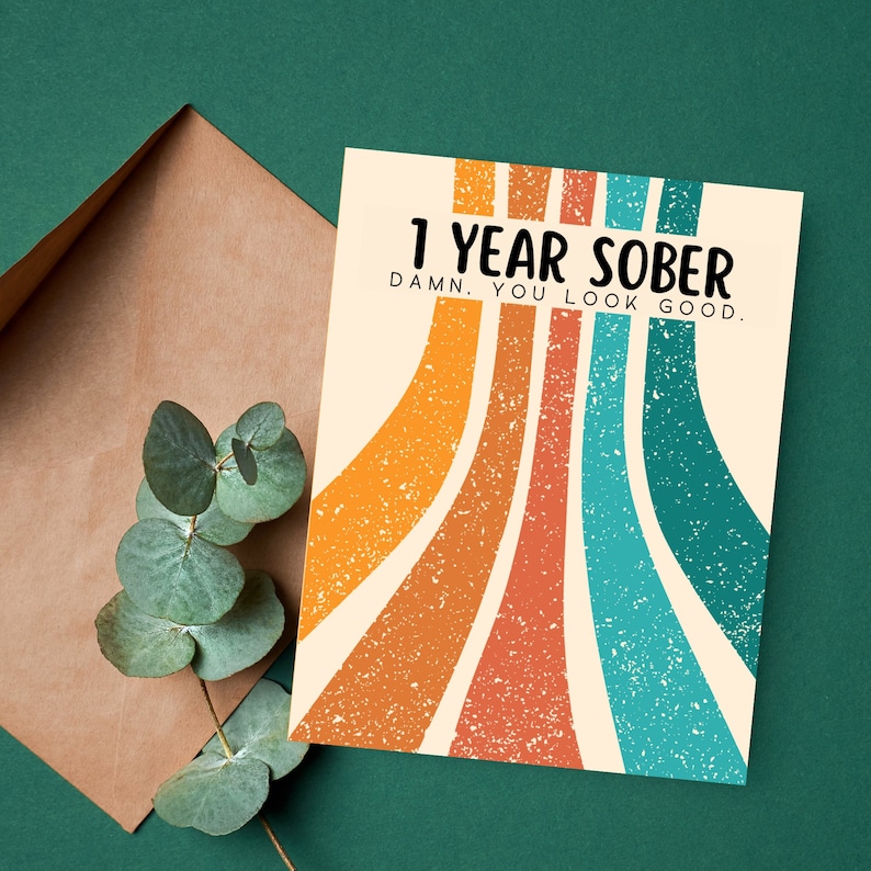 May include: A card with a retro design featuring colorful stripes and the text "1 Year Sober. Damn. You look good."