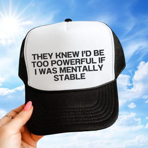 May include: A black and white trucker hat featuring the text "THEY KNEW I'D BE TOO POWERFUL IF I WAS MENTALLY STABLE". The hat has a white front panel and a black mesh back, with a curved peak.