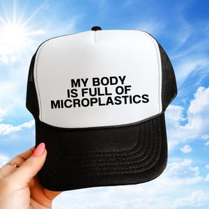 May include: A black and white trucker hat with the text "MY BODY IS FULL OF MICROPLASTICS" in bold, black letters. The hat has a white front panel and a black mesh back. The hat is worn by a person.