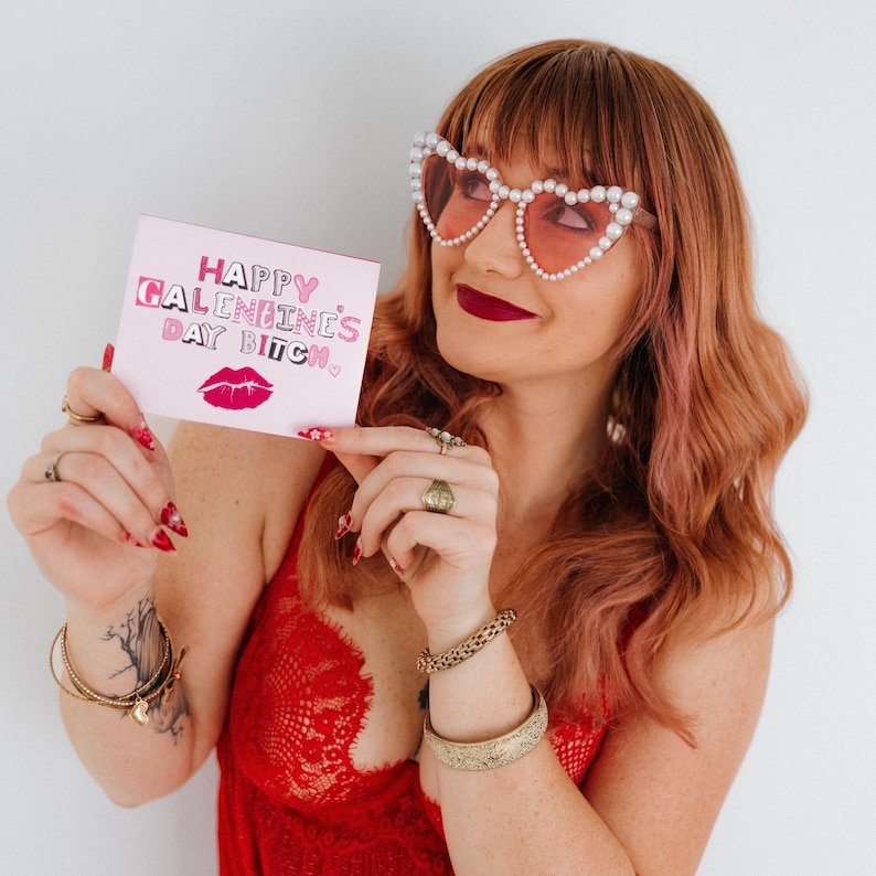 May include: A pink card with the text "Happy Galentine's Day Bitch" in pink and white letters with a pink lip print below the text. A person is holding the card in front of their face, wearing heart-shaped sunglasses with pearl accents.