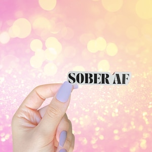 May include: A hand holding a white card with the text "SOBER AF" in black letters. The background is a pink and yellow bokeh.
