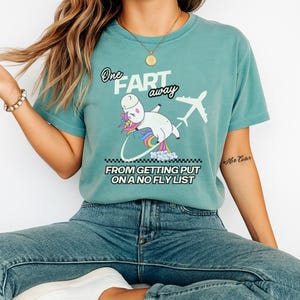 Op de afbeelding: Een blauwgroen t-shirt met korte mouwen en een afbeelding van een eenhoorn met een regenboogkleurige manen en staart. De tekst luidt One FART away FROM GETTING PUT ON A NO FLY LIST, met een wit vliegtuigsilhouet en een wolkenspoor.