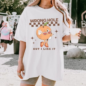 May include: White t-shirt with the text "WRONG HOLE" above a cartoon orange wearing sunglasses and the text "BUT I LIKE IT" below. The orange has arms, legs, and is wearing shoes. The shirt is a relaxed fit.