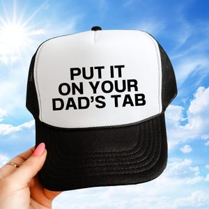 May include: Black and white trucker hat with the text "PUT IT ON YOUR DAD'S TAB" in bold, black letters. The hat has a white front panel and a black mesh back. The hat is being held up against a blue sky.