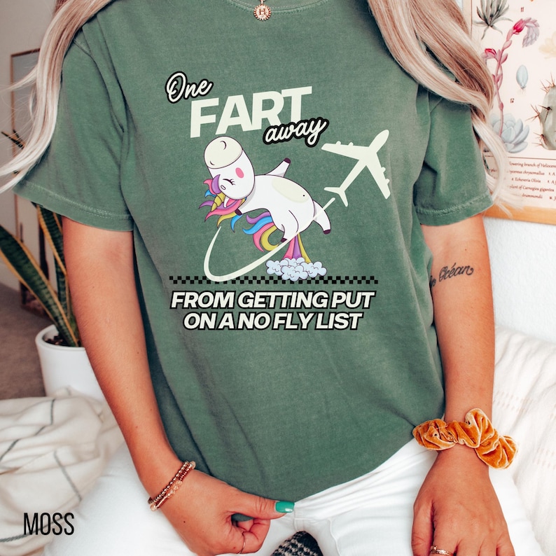 Op de afbeelding: Een mosgroen t-shirt met een afbeelding van een witte eenhoorn met een regenboogkleurige manen en staart. De eenhoorn vliegt naast een vliegtuig en laat een spoor van wolken achter. De tekst luidt: One FART away FROM GETTING PUT ON A NO FLY LIST.