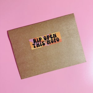 Puede incluir: Un sobre de papel marr&oacute;n con una pegatina colorida que dice "RIP OPEN THIS MOFO" en texto negro.