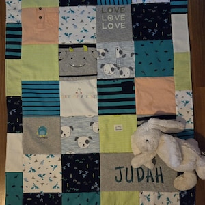 Op de afbeelding: Een patchworkdeken gemaakt van verschillende kindershirts, met afbeeldingen van dinosaurussen, puppy's en monsters. De quilt bevat de naam "Judah" geborduurd in grijs en een witte pluchen konijn.