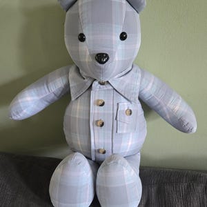 May include: A stuffed teddy bear crafted from light blue and white plaid fabric. The bear features a collared shirt front with four buttons, a small chest pocket, two black circular eyes, and a black nose. The limbs and ears are made of the same plaid material.