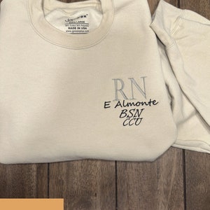 May include: Cream-coloured sweatshirt with embroidered text. The text includes "RN", "E Almonte", "BSN", and "CCU" in grey and black thread. The sweatshirt is folded on a wooden surface. The brand name "GOWEN" is visible.