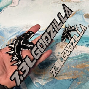 May include: Black and silver 7.3L Godzilla emblem featuring a Godzilla illustration with red eyes. The emblem has a 3D effect and is designed with a stylized font. The image shows two emblems, one held in a hand and another on a surface.