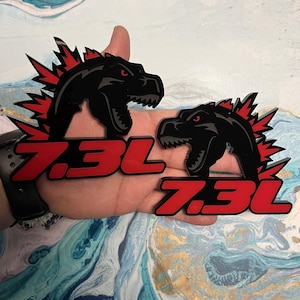 May include: Two black and red acrylic decals featuring a stylized dinosaur head with a red mohawk and the text "7.3L".