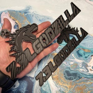 May include: Black and gray 7.3L GODZILLA emblem with a carbon fiber pattern. The emblem features a Godzilla silhouette and the text "7.3L GODZILLA". The design is three-dimensional and appears to be made of a durable material.