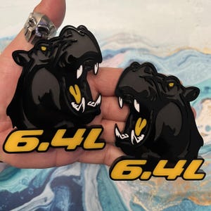 May include: Two black and yellow 6.4L emblems featuring a cartoon black panther with a white mouth and yellow teeth. The emblems are made of a glossy material.