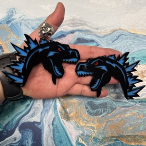 May include: Two black and blue dinosaur head pins with red eyes and a spiky blue crest. The pins are on a hand.