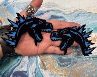 May include: Two black and blue dinosaur head pins with red eyes and a spiky blue crest. The pins are on a hand.
