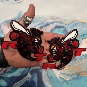 May include: Two red and black 3D car emblems with a stylized bee design and the letters "RT" in red. The bee has white wings and yellow eyes.